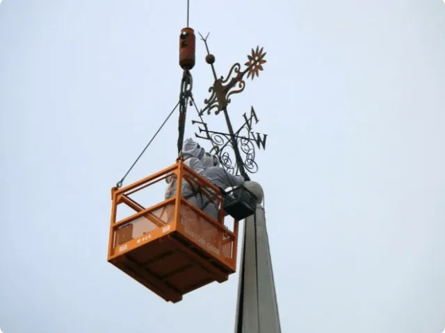 two people in a suspended container reaching for a weather vane
