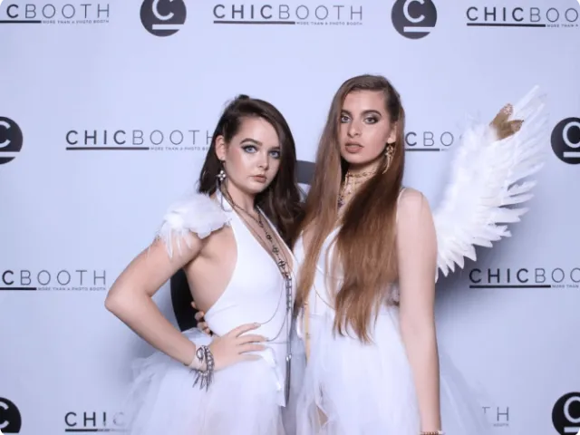 two women dressed in all white posing in front of a branded backdrop