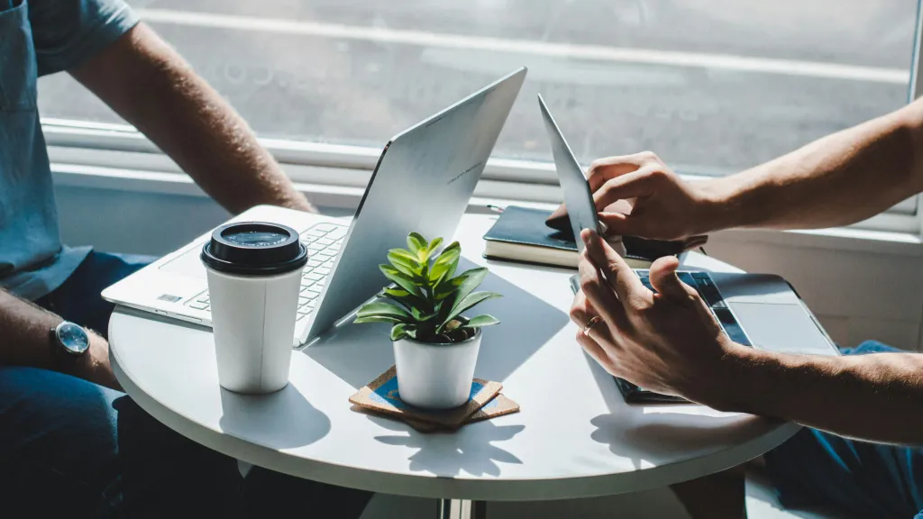 Two people sit across from each other at a small round table on their laptops. Only their forearms are visible.