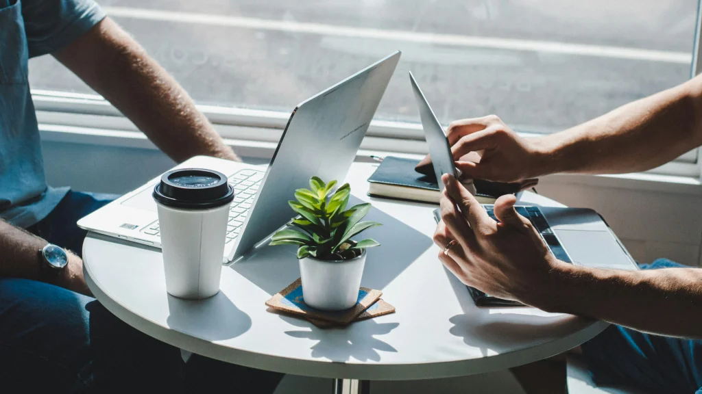 Two people sit across from each other at a small round table on their laptops. Only their forearms are visible.