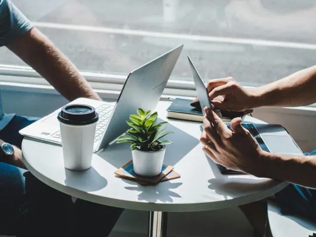 Two people sit across from each other at a small round table on their laptops. Only their forearms are visible.