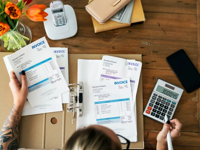 A person sits at their desk with invoices and a calculator