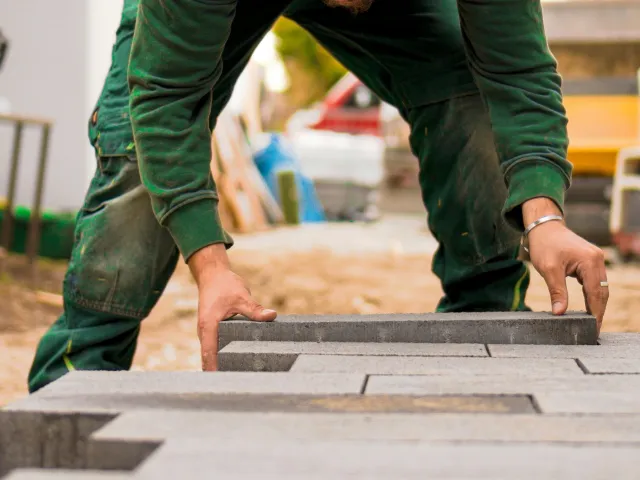 A person in a green shirt lays a brick on the ground, building a floor