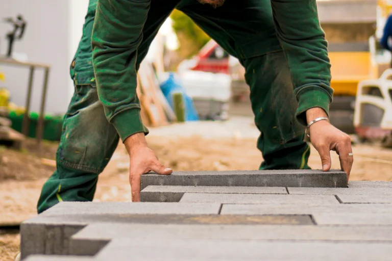 A person in a green shirt lays a brick on the ground, building a floor