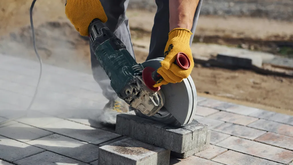 A pair of hands in work gloves hold a circular saw to cut a brick