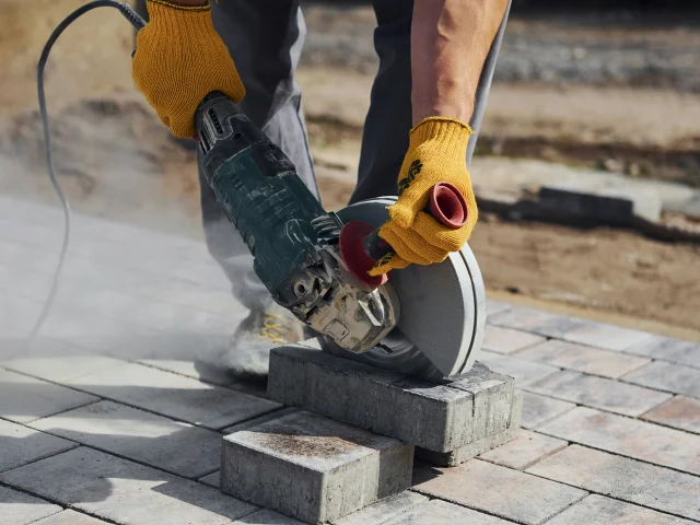 A pair of hands in work gloves hold a circular saw to cut a brick