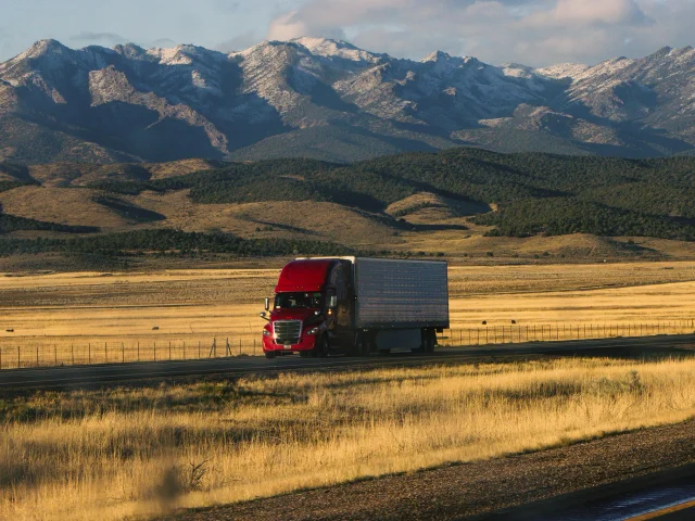 A truck drives down a road with snow-capped mountains in the background