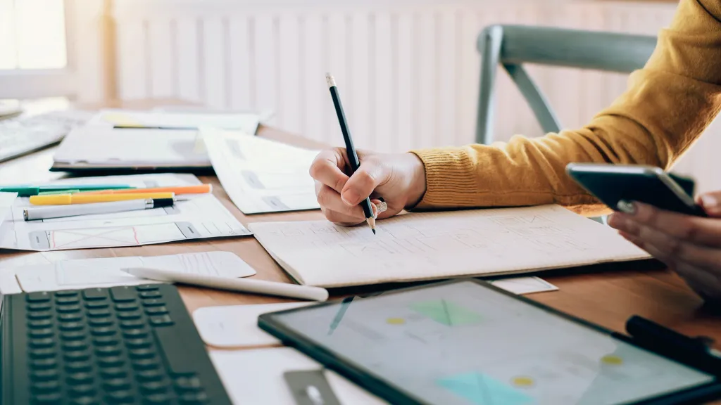 Hand writing on notebook on a desk filled with paperwork and a keyboard