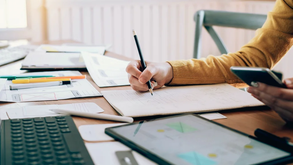 Hand writing on notebook on a desk filled with paperwork and a keyboard