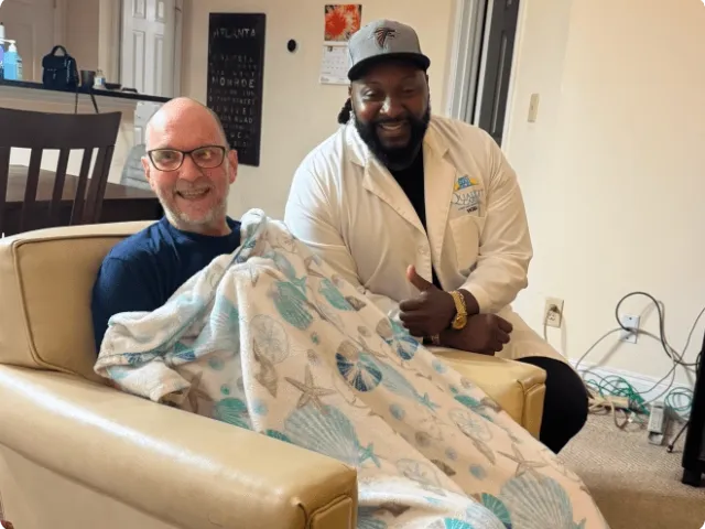 medical professional next to an old man in a chair with a blanket draped over him, both people smiling at the camera