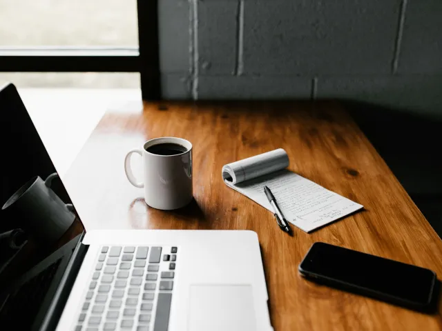 Wooden desk against a windowed wall with laptop, coffee, notepad