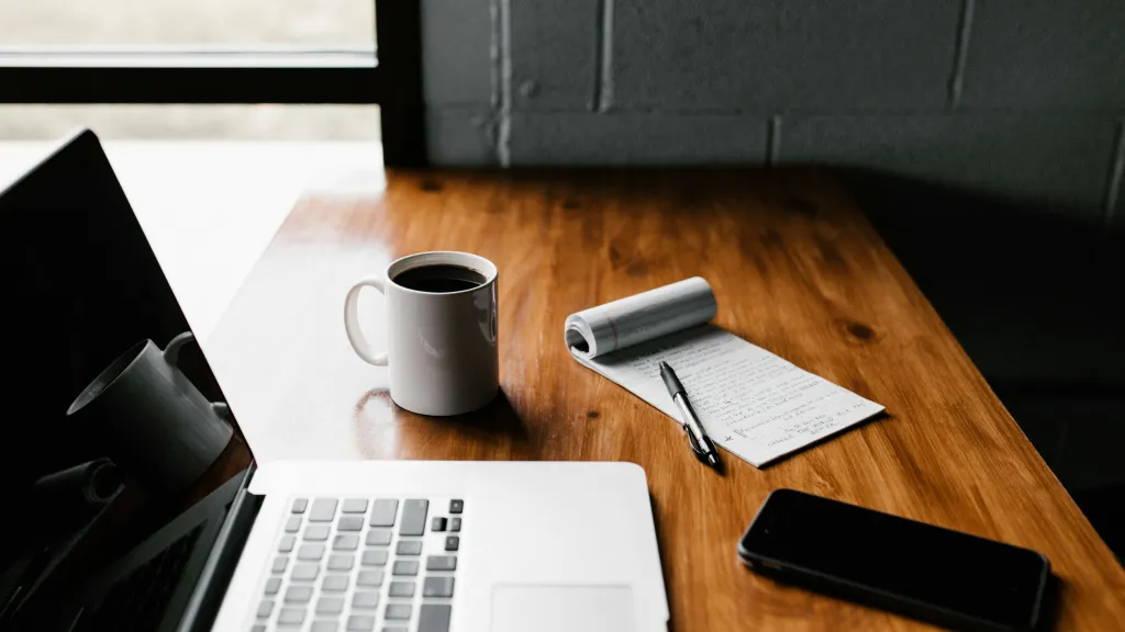 Wooden desk against a windowed wall with laptop, coffee, notepad