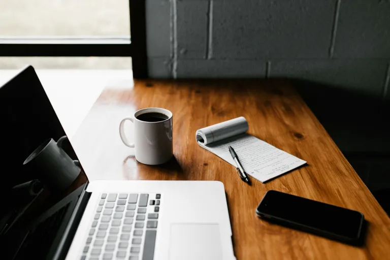 Wooden desk against a windowed wall with laptop, coffee, notepad