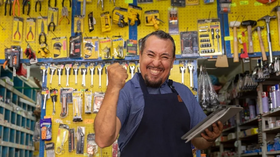 man with dark hair and mustache wearing apron standing in front of a wall of tools smiling and pumping his fist to illustrate article about starting a home services business