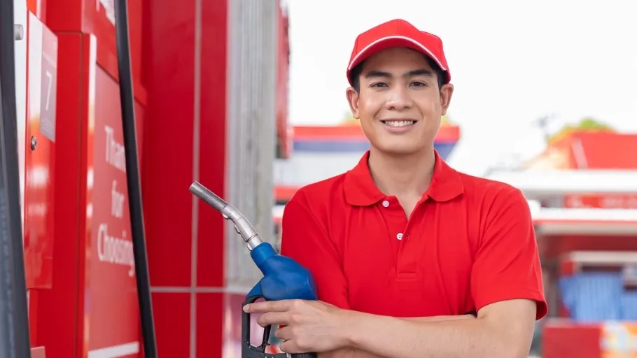 Young man with dark hair wearing red had and shirt standing at gas pump smiling to illustrate article about best fleet cards