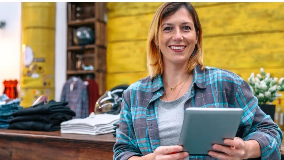 Smiling woman with blonde hair wearing a plaid shirt and holding a tablet in a clothing store to illustrate article about starting a business with no money,