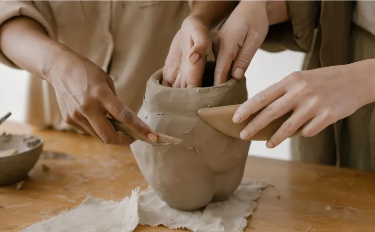 Close-up of two artisans sculpting a clay vase together in a pottery studio.