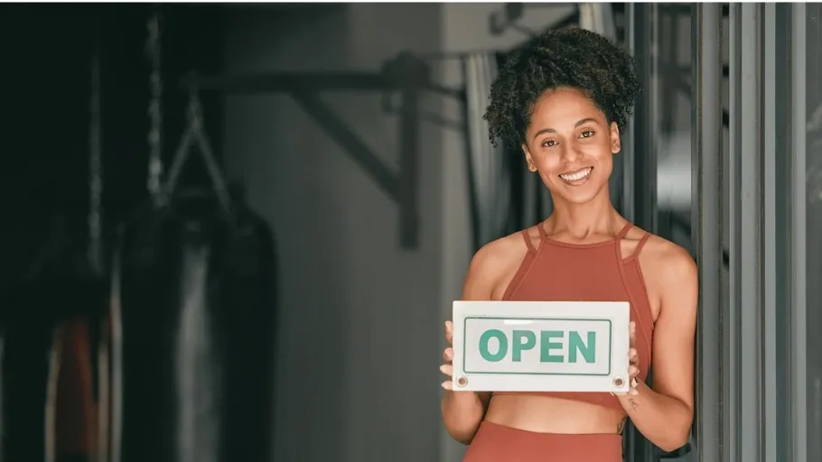 Younger woman with dark hair in a pony tail wearing orange fitness clothing, standing in front of fitness equipment and holding a sign that says open to illustrate article about cheapest franchises