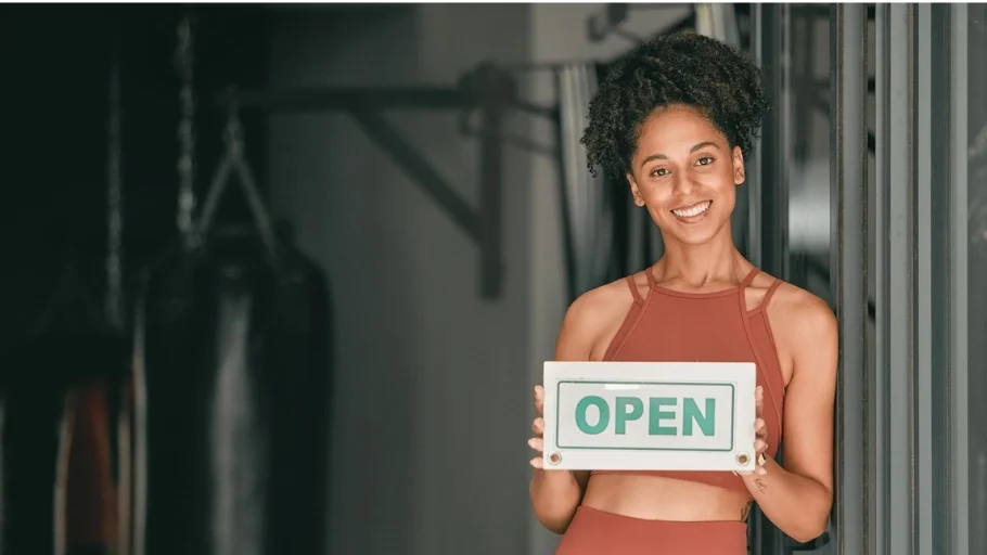 Younger woman with dark hair in a pony tail wearing orange fitness clothing, standing in front of fitness equipment and holding a sign that says open to illustrate article about cheapest franchises