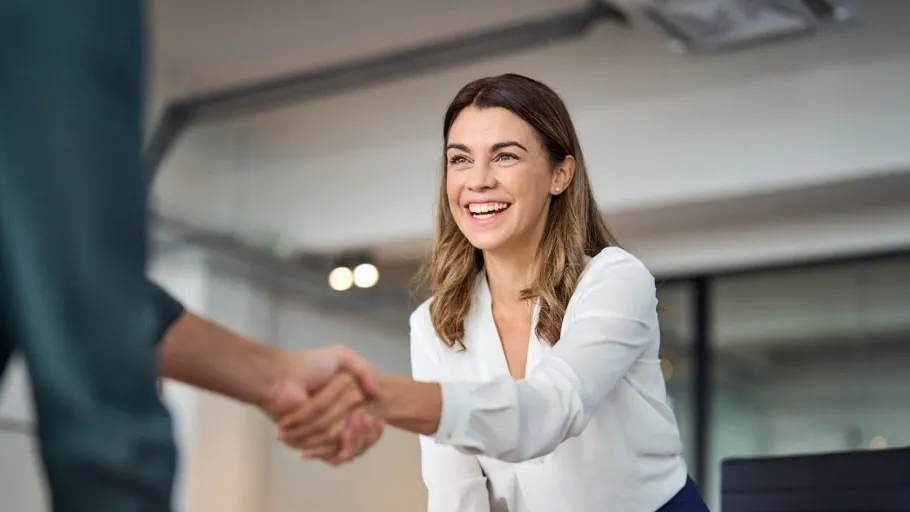 Woman with brown hear wearing a white top, leaning across table shaking hands with someone to illustrate article about business broker fees.