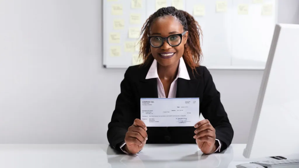 Woman with braided hair wearing dark suit and glasses holding a check and smiling to illustrate article about paying yourself as a business owner
