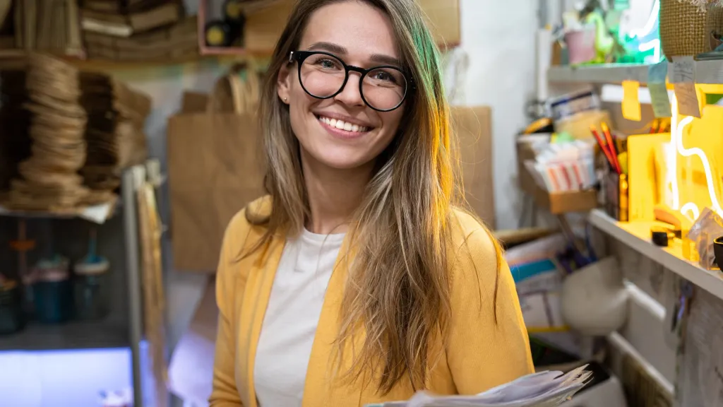 A female business owner with long hair and glasses in a yellow sweater holding paperwork and smiling to illustrate paying California franchise tax
