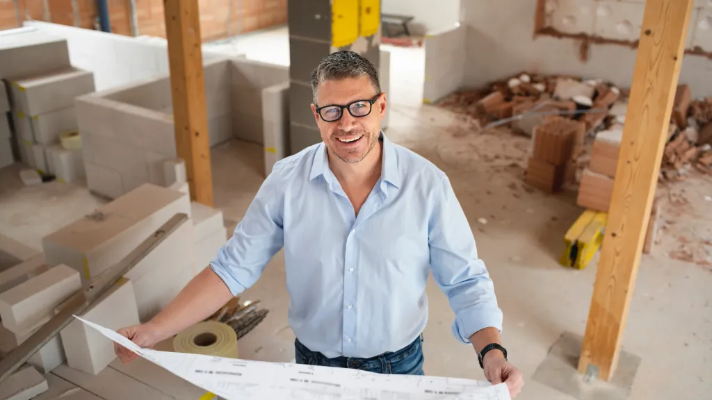 man with black glasses and blue shirt checking plans and construction progress on building site used to illustrate article about high risk business loans