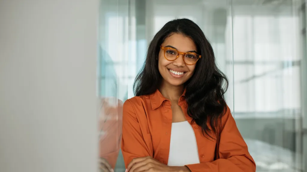 Young female professional with long hair and orange jacket standing smiling at camera to illustrate article about how subscriptions help build credit