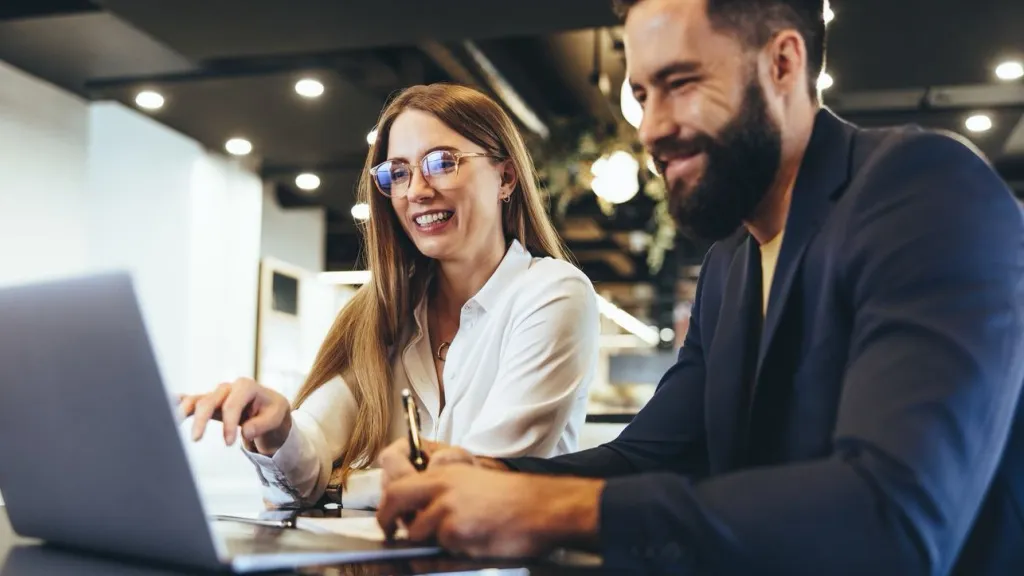 Happy young entrepreneurs smiling. Woman with long hair and glasses and man with beard working together on laptop in a modern workspace to illustrate article about small business coaches and mentors.