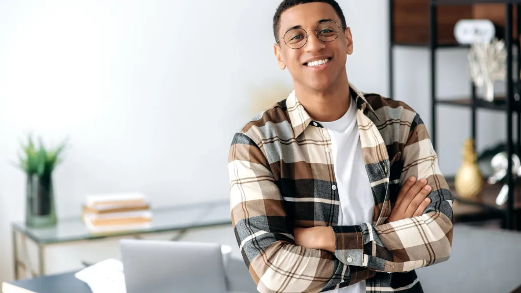 Successful confident young man with glasses, stylishly dressed, standing near work desk with arms crossed, looking at camera with friendly smile to illustrate article about business life stages.