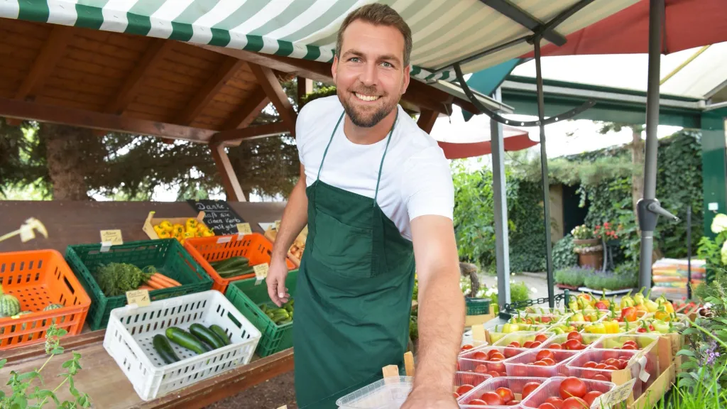 Business owner male in apron smiling at camera selling fruits and vegetables at farmers market to illustrate article about building business credit