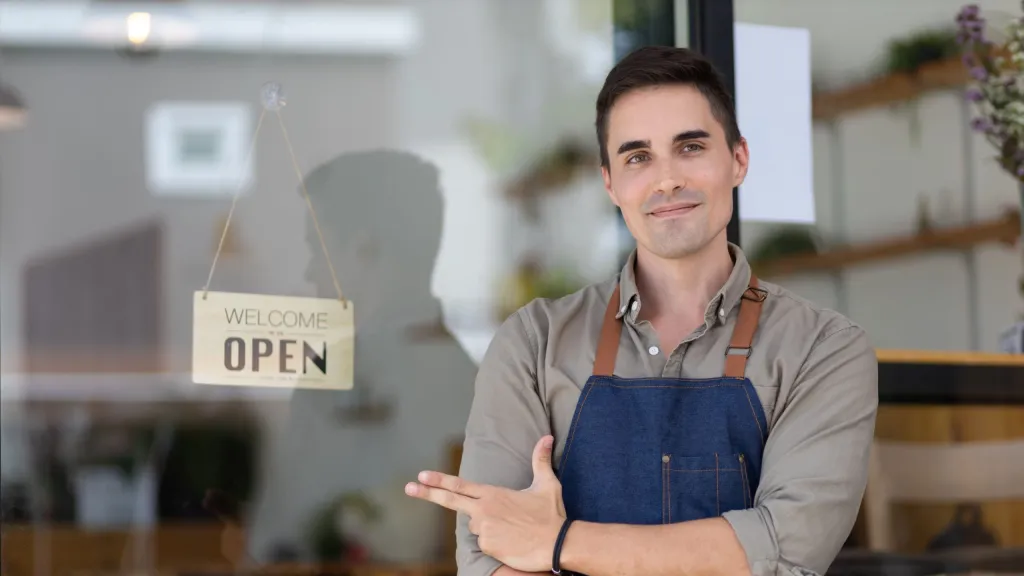 Man with dark short hair wearing blue apron and brown shirt pointing to and Open sign to illustrate article about financing and grants for Native American owned business.