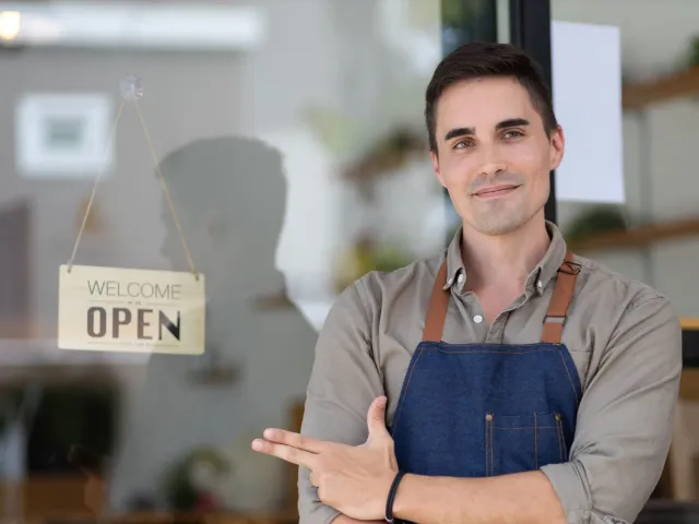 Man with dark short hair wearing blue apron and brown shirt pointing to and Open sign to illustrate article about financing and grants for Native American owned business.
