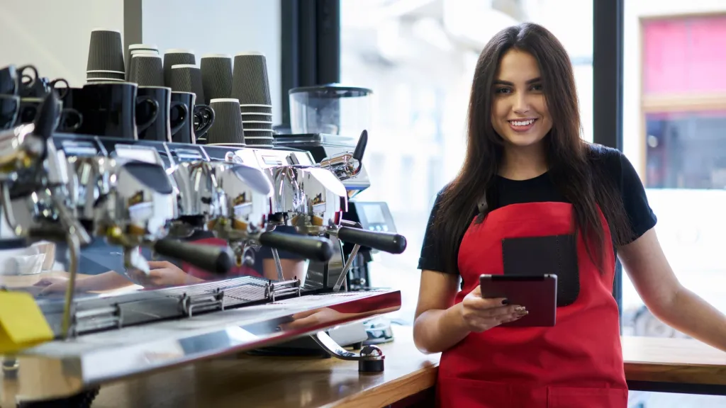 Smiling woman with long dark hair wearing a red apron standing by expresso bar with tablet in hand used to illustrate article about highest possible business credit scores