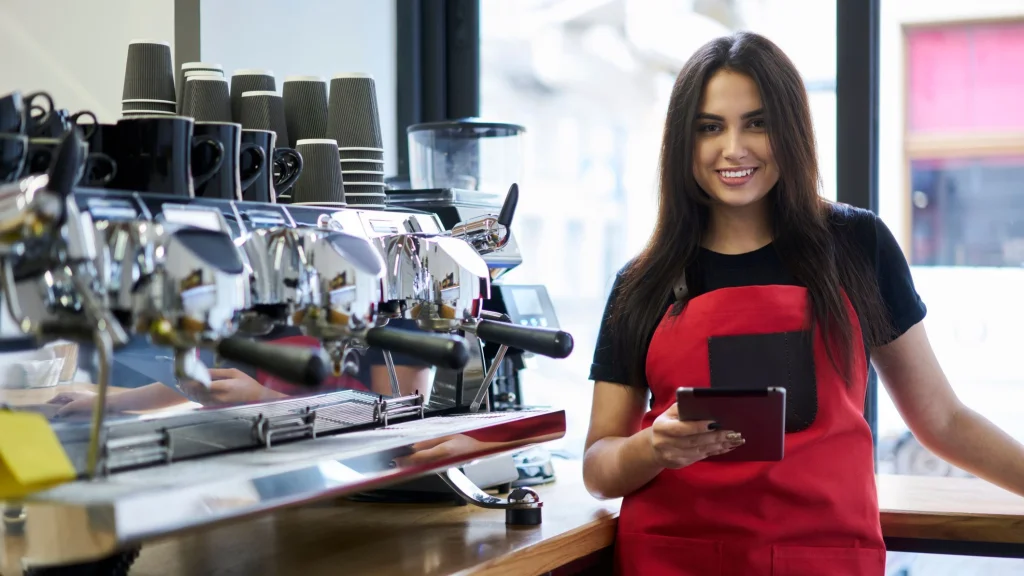 Smiling woman with long dark hair wearing a red apron standing by expresso bar with tablet in hand used to illustrate article about highest possible business credit scores