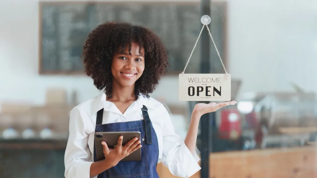 Young woman with black curly hair smiling wearing apron and holding open store at cafe