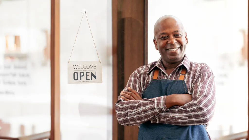 Man wearing Apron and checkered short smiling and standing in front of door with open sign to illustrate article about Small Business Saturday.