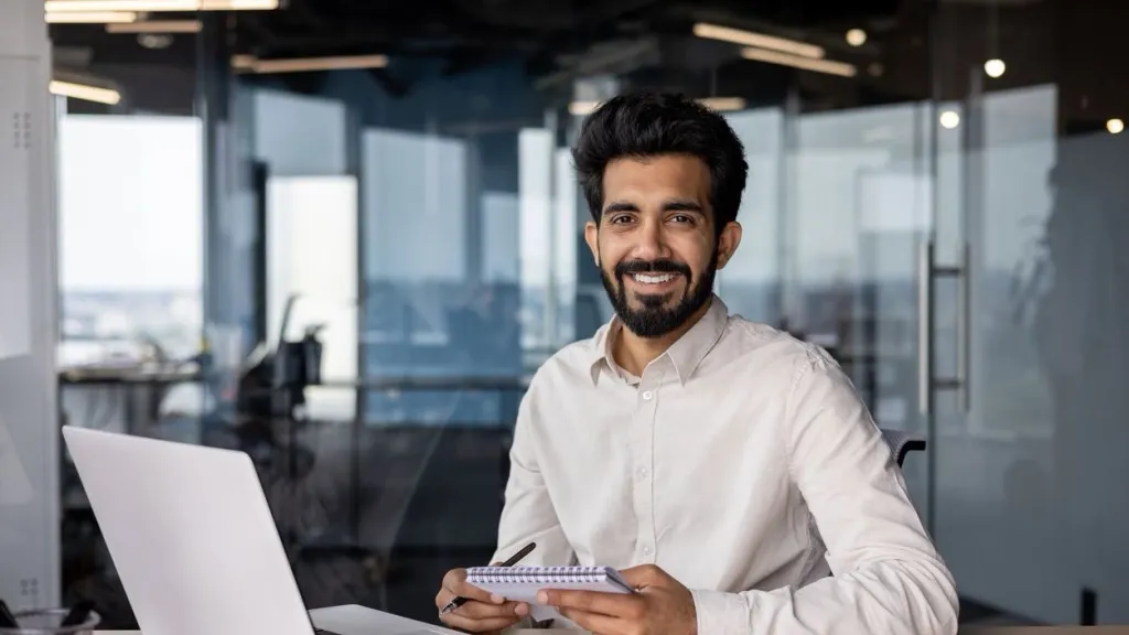 Younger Indian man smiling and looking at the camera, sitting in the office at a desk with a laptop, holding a notebook to illustrate article about tax write offs.