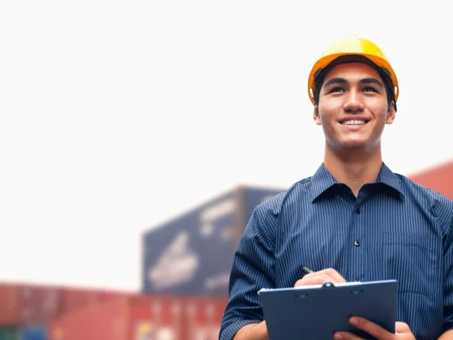 Smiling man wearing a yellow hard hat and blue shirt and holding a clipboard, standing in front of shipping containers to illustrate article about tariffs and small business owners.