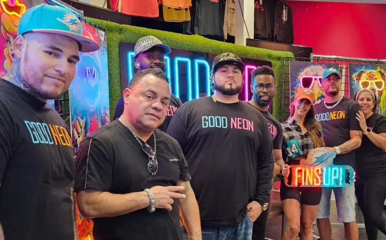GoodNeon team posing together inside their neon sign and custom apparel shop surrounded by colorful artwork and illuminated signs.