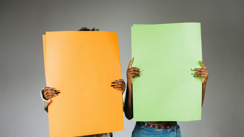 Two people hold up two different brightly colored pieces of paper in front of their faces and torsos