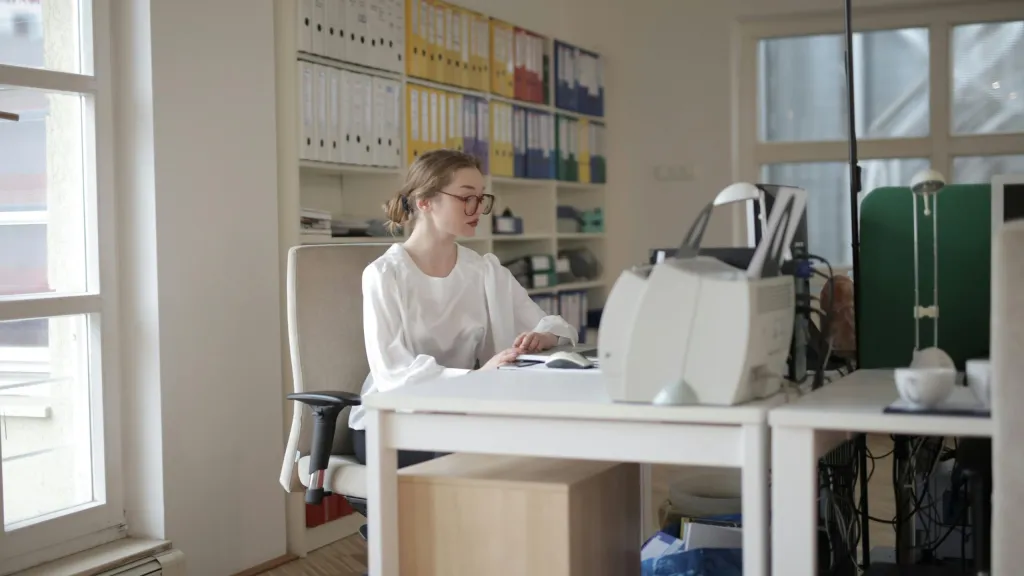 Young woman with brown hair tied back, wearing glasses and a long sleeved white shirt sitting at computer to illustrate article about bookkeeping 101