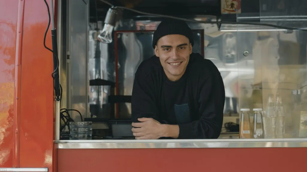 Man smiling looking at camera through food truck window wearing a dark turtleneck and dark hat, to illustrate article about starting a business without loans
