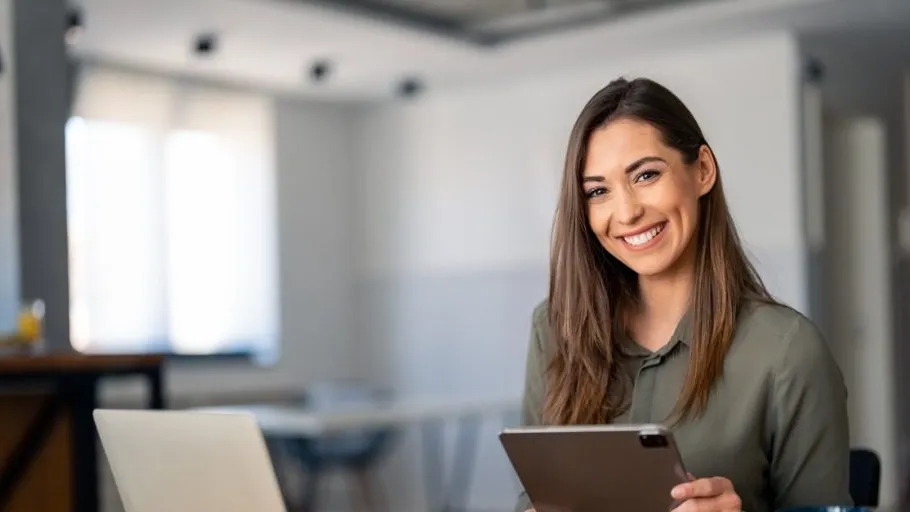 Younger woman with long brown hair in office smiling and holding tablet to illustrate article about FICO vs business credit scores