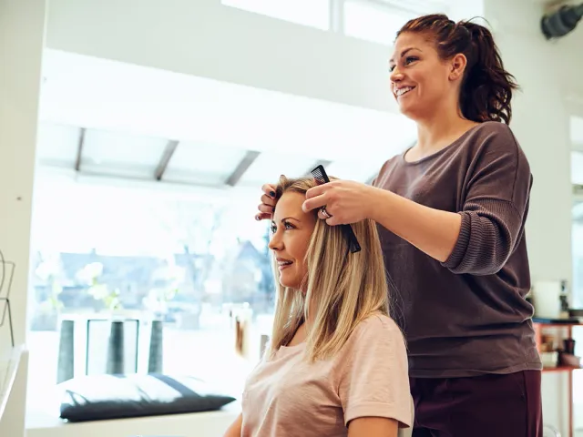 Hairdresser smiling while styling a client’s hair in a bright, modern salon with large windows and hair products on display
