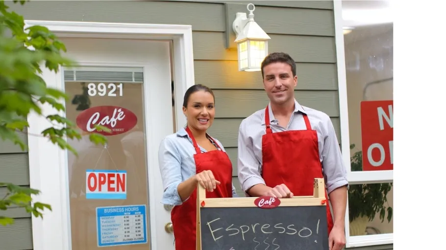 man and woman both with brown hair wearing aprons standing in front of cafe with chalk board sign to illustrate article about small business loan requirements