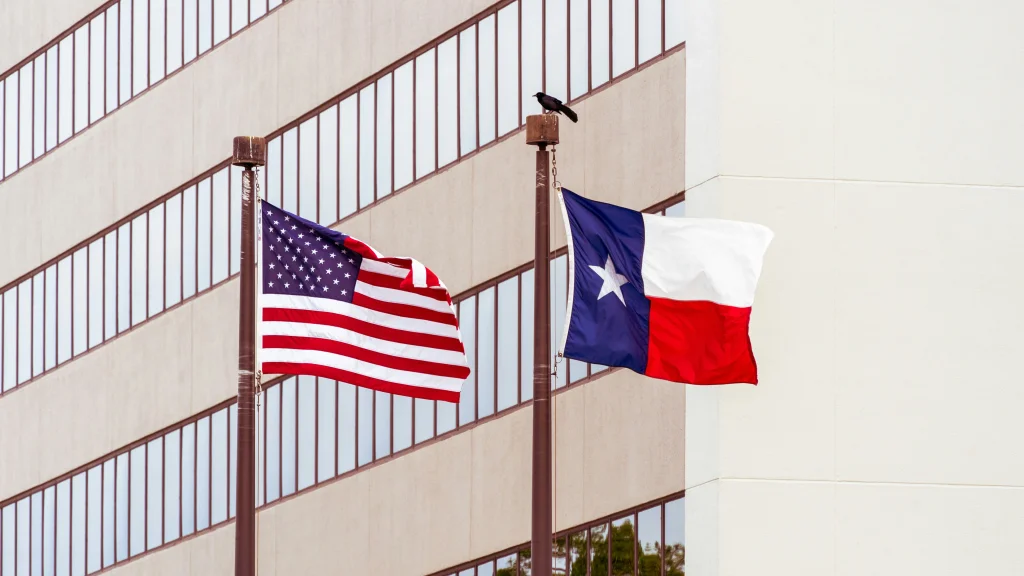 An American flag and Texas state flag wave in the breeze with an office building behind them