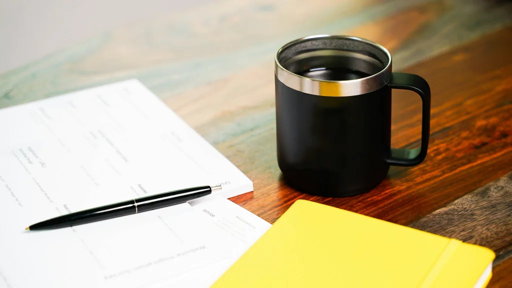 A pen, a paper, a pad and a coffee cup on a desk. Perhaps the person is comparing the types of cyber insurance.