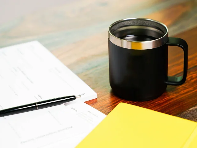 A pen, a paper, a pad and a coffee cup on a desk. Perhaps the person is comparing the types of cyber insurance.