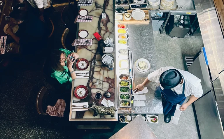 Overhead view of a restaurant kitchen and dining counter with staff preparing food and a customer seated.